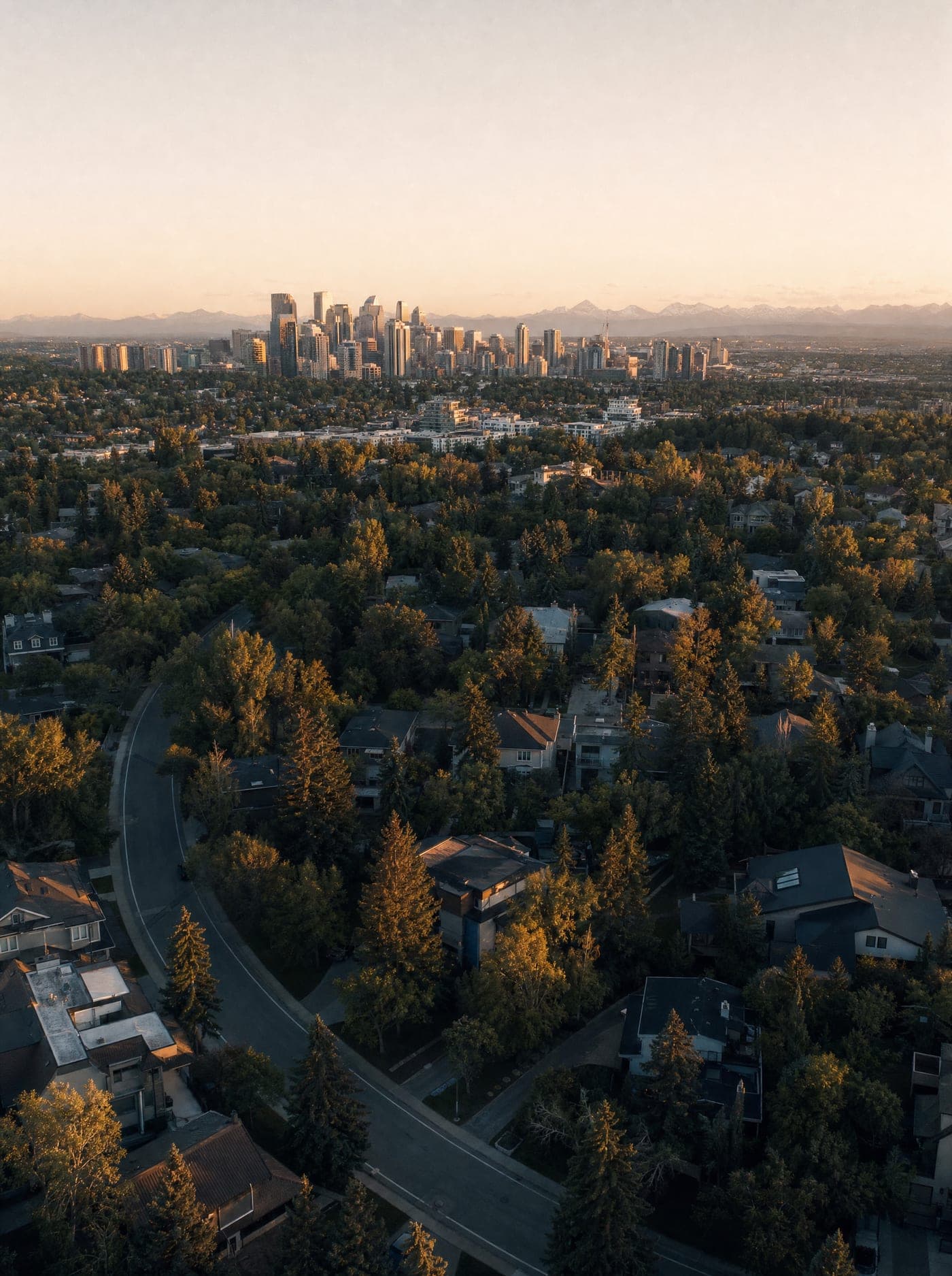 Golden-hour aerial view over a southwest Calgary neighbourhood, the downtown skyline and the Rockies on the horizon.
